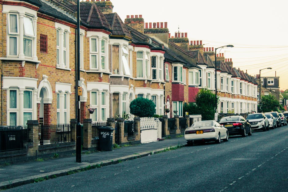 A quiet residential street featuring a row of red brick terraced houses with pitched roofs and chimneys, surrounded by trees with bare branches indicating late autumn or early winter. The narrow aspha