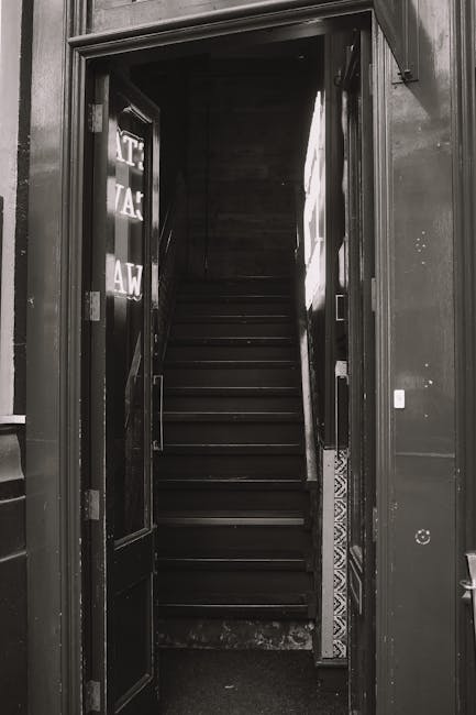 A metal staircase with narrow, evenly spaced steps and a handrail on one side, positioned between two brick walls and a concrete wall in an outdoor setting. The staircase leads up to a small platform