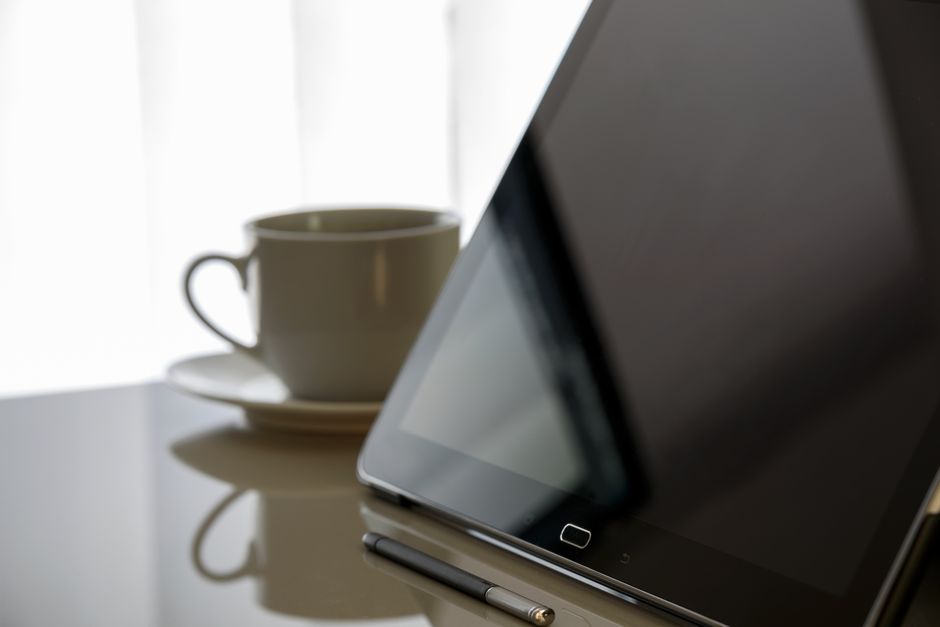 A close-up image showing a white ceramic teacup and saucer positioned on a reflective surface, with a dark tablet device resting at an angle nearby, accompanied by a metallic stylus pen. The backgroun