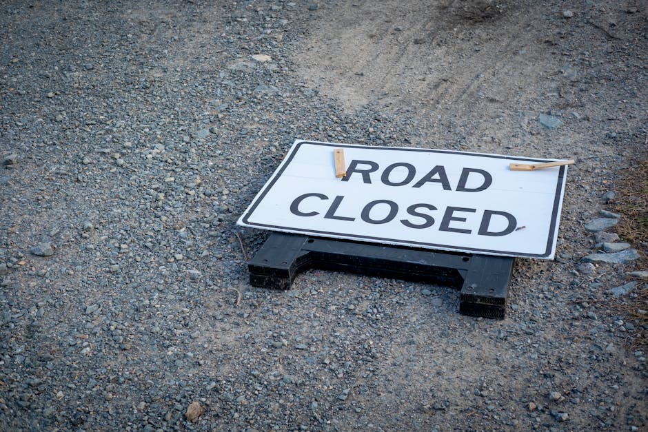 A daytime outdoor scene showing a narrow sidewalk section temporarily closed for a home relocation or moving process, with the pathway blocked by orange and white traffic cones and a rectangular orang