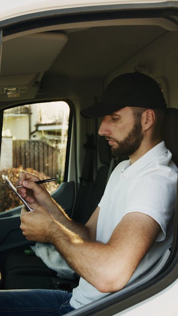 A man in casual clothing is standing outside a residential property next to a large white delivery van with an open rear door, engaged in a home relocation or furniture transport process. He is liftin
