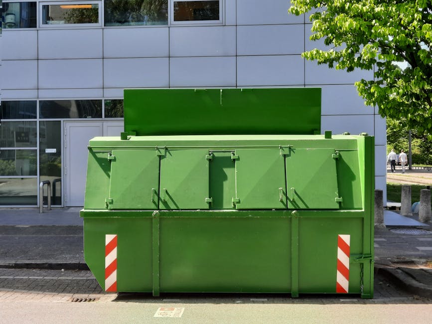 An outdoor scene featuring a large, metal waste dumpster with a curved lid, positioned on a pavement next to a brick wall with weathered mortar and greenery growing over the top. The dumpster appears 
